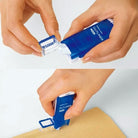 Hands opening and applying a patch of adhesive with Blue Kokuyo Dotliner Adhesive Roller Stamp with white logo onto brown paper