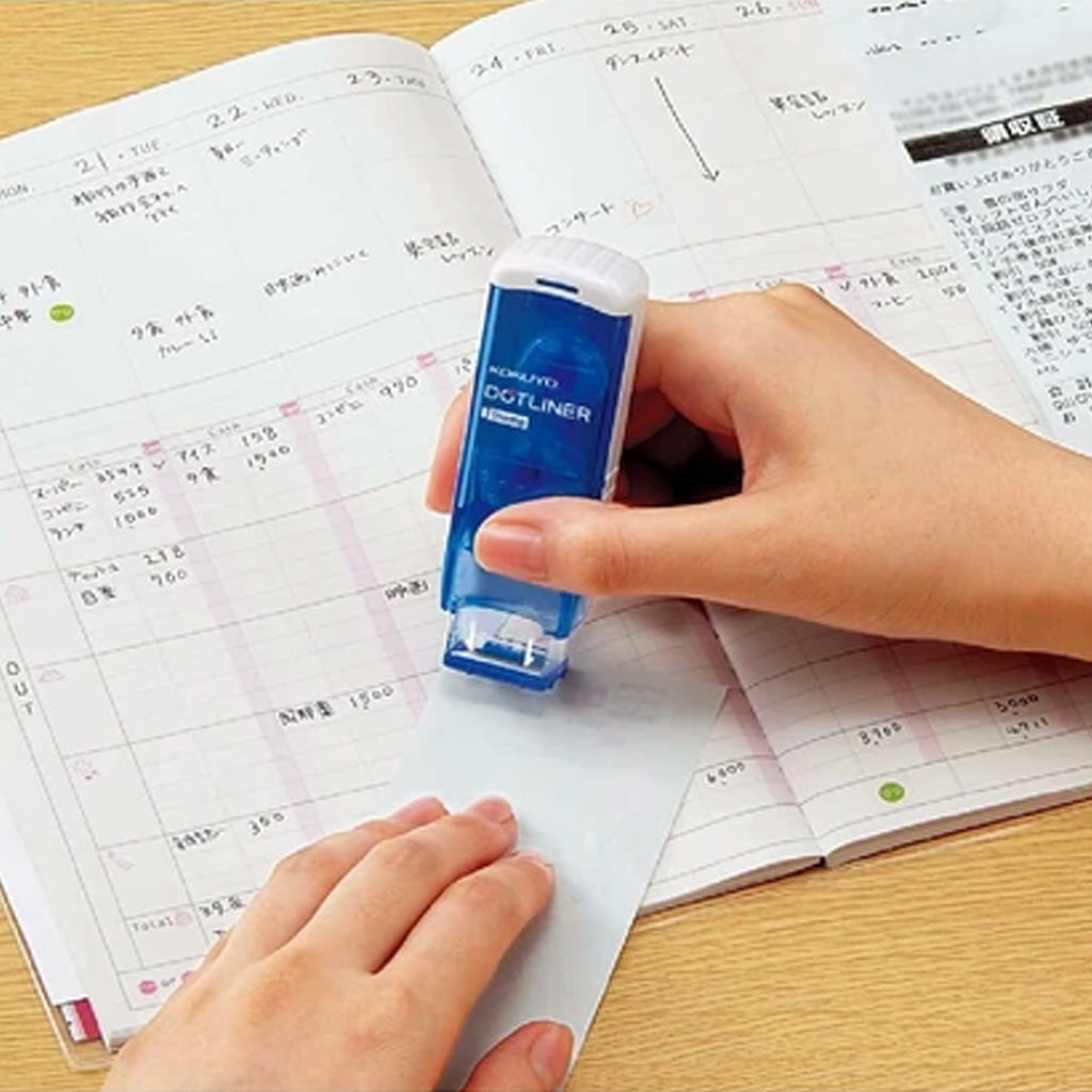 A person's hand holding a Blue Kokuyo Dotliner Adhesive Roller Stamp with white logo and applying a patch of glue onto strip of paper in front of planner with Japanese writing