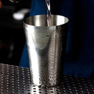 Metal cocktail shaker on a bar surface with liquid being poured into it.