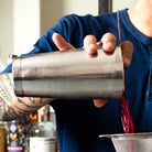 Person holding a metal cocktail shaker with a blurred bar setting in the background