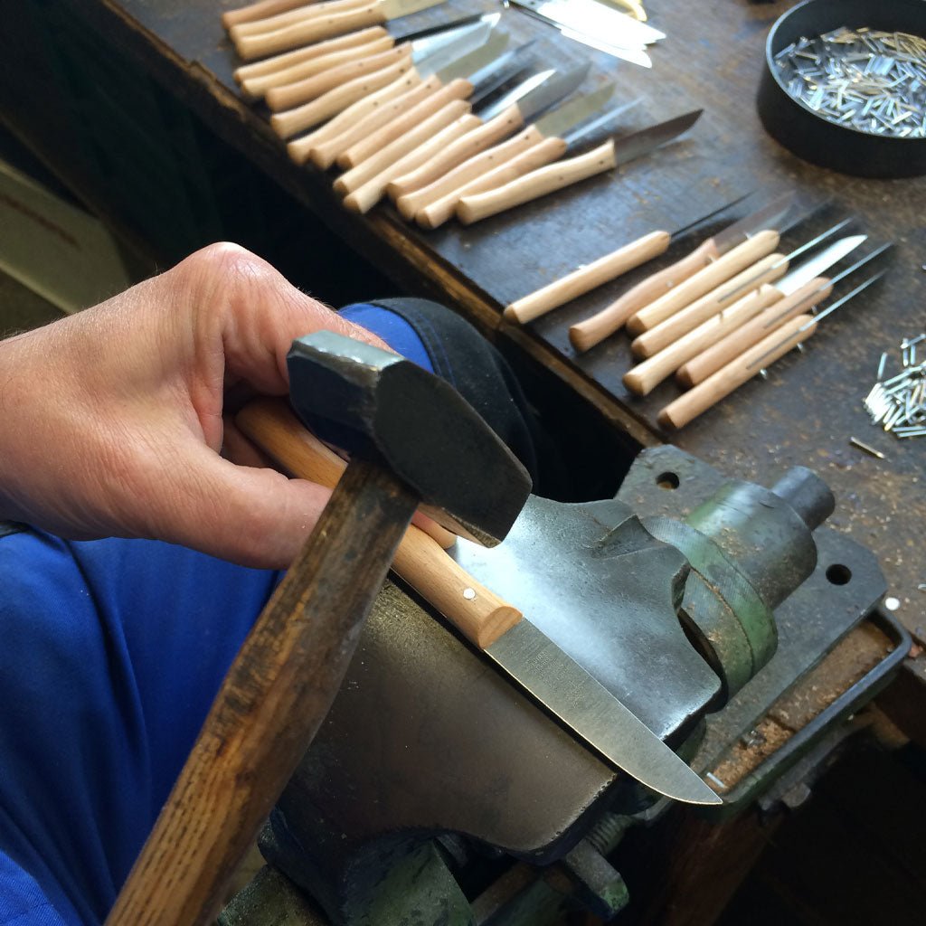 Hand holding a hammer over a knife with a wooden handle on a workbench with tools in the background