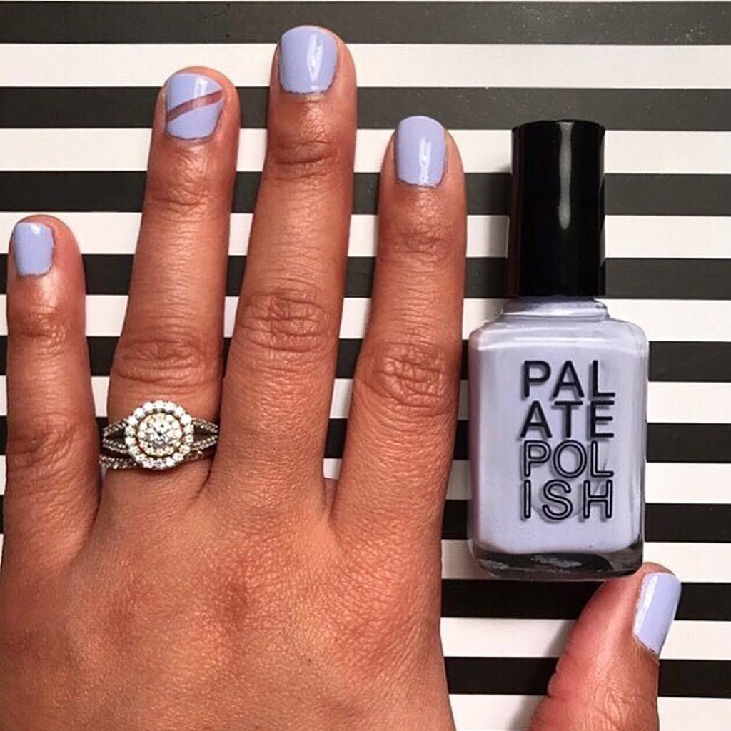 Hand with light purple nail polish holding a bottle of 'Palate Polish' against a black and white striped background.