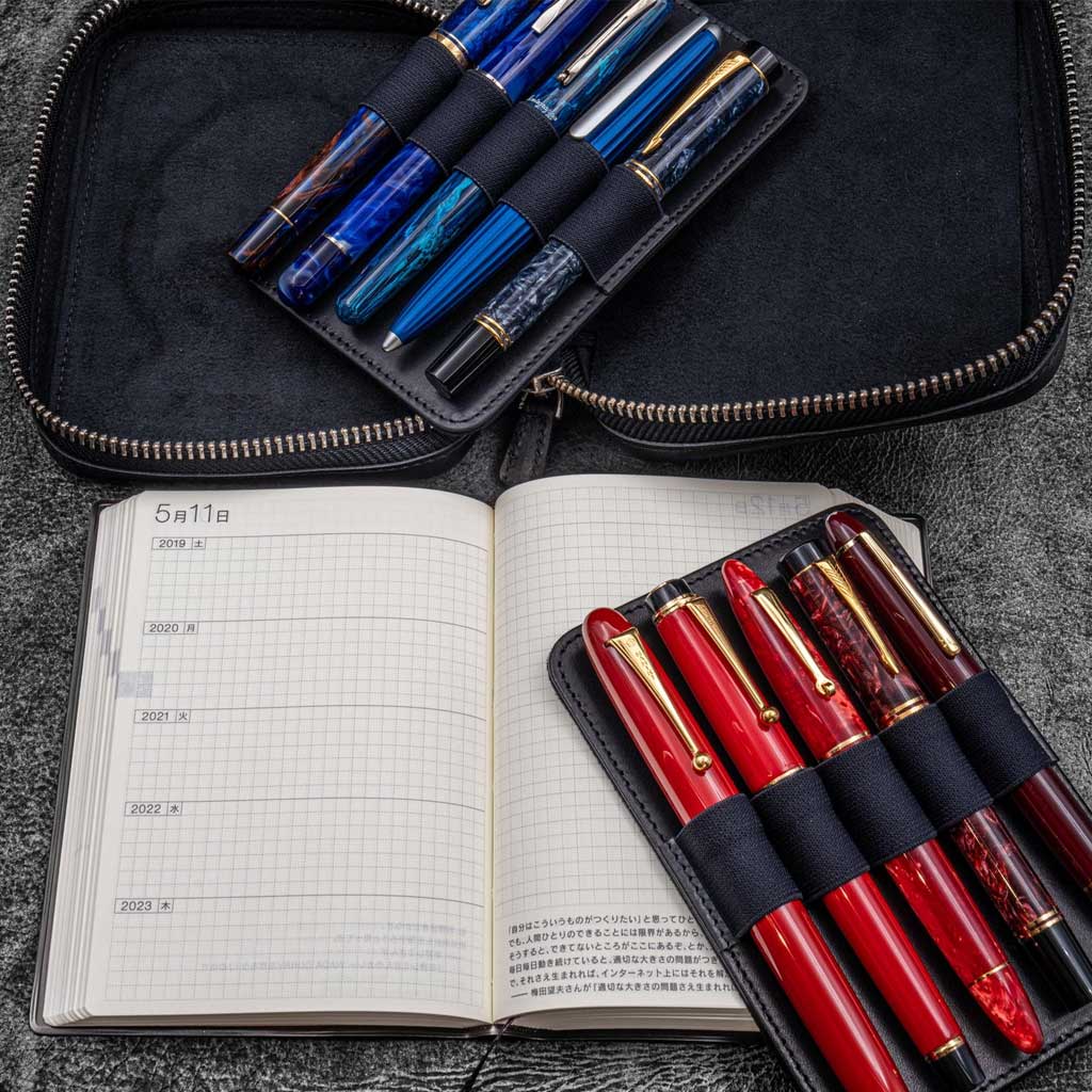 Black leather zippered pen pouch with removable holders for ten pens and open planner notebook on a dark surface