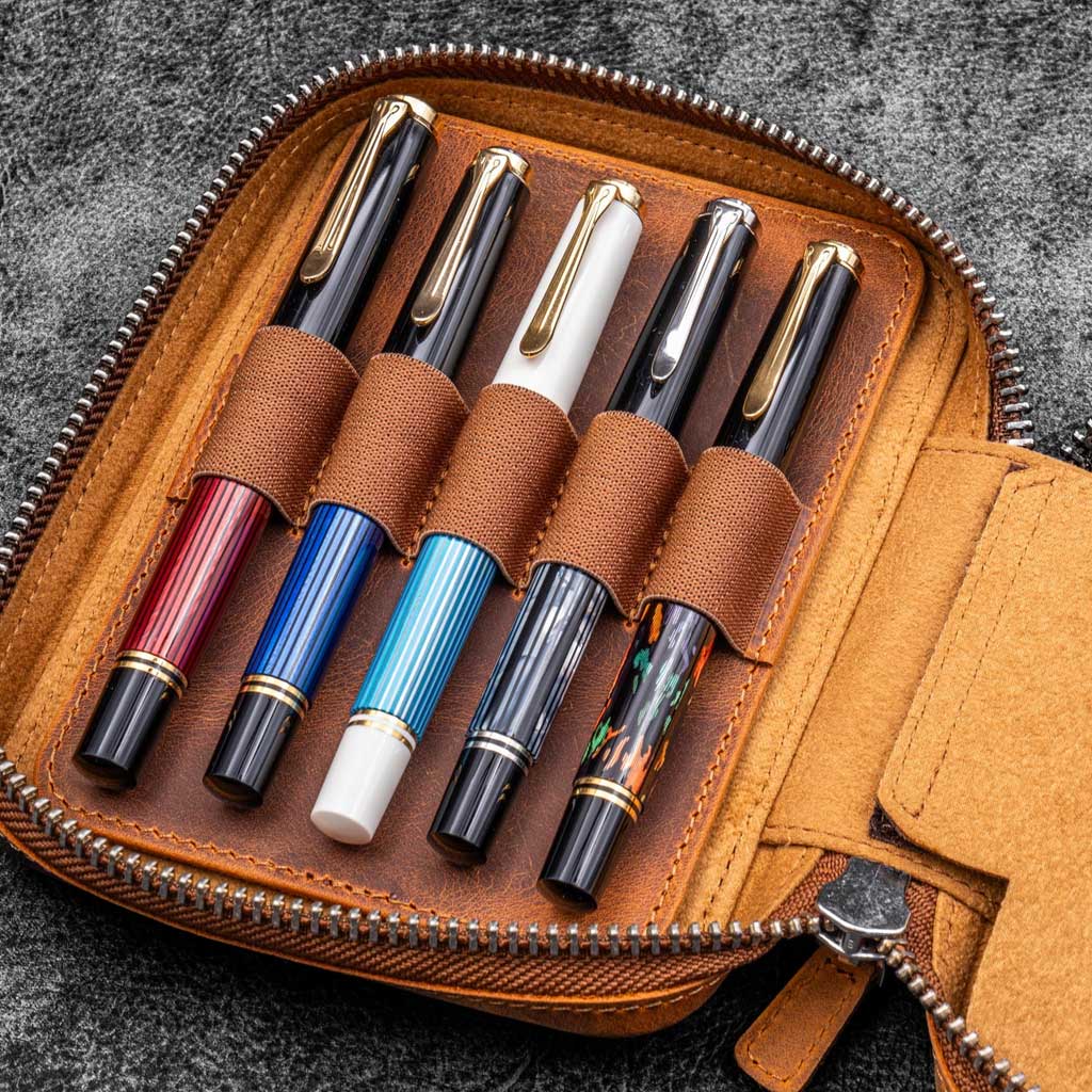 Brown leather zippered pen pouch with five pens on a dark surface