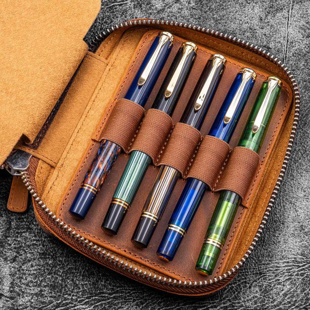 Brown leather zippered pen pouch with five pens on a dark surface
