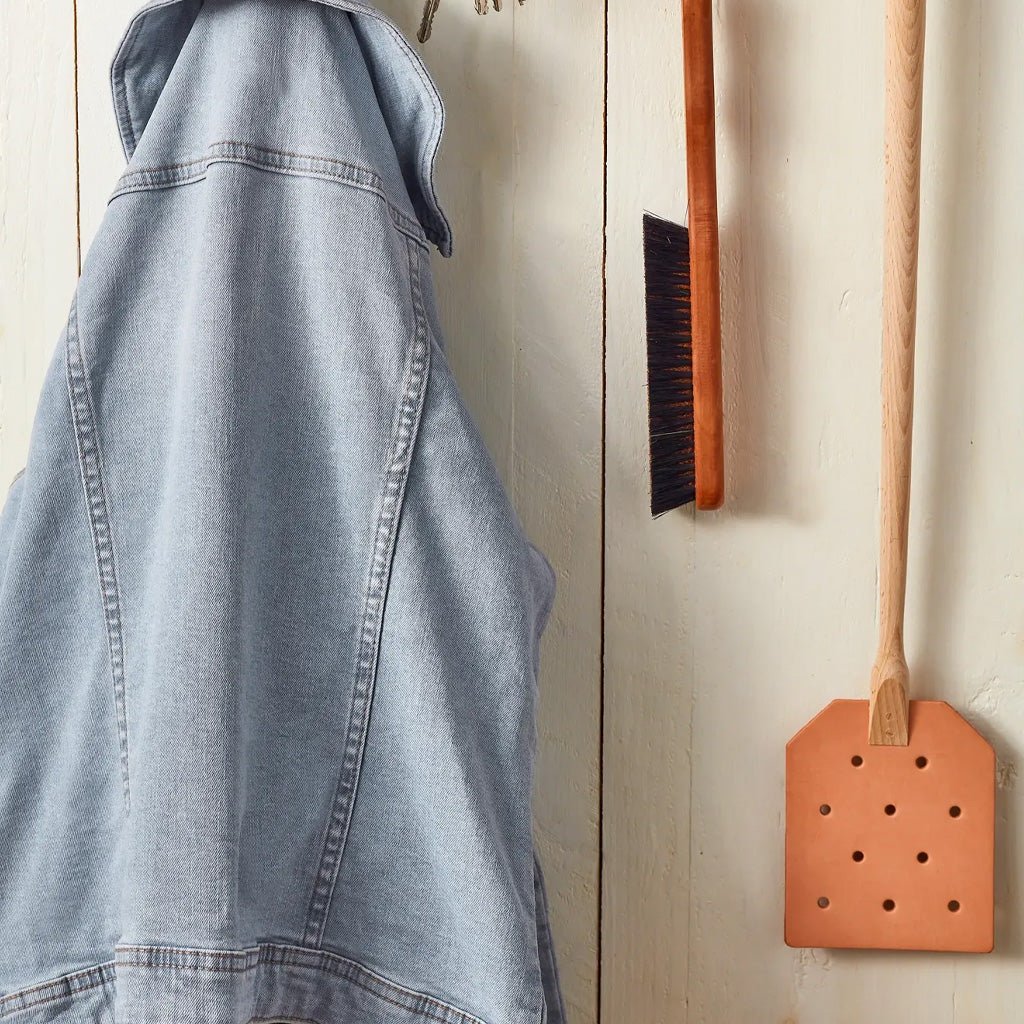 A light blue denim jacket hangs on a cream-colored wooden wall next to a wooden fly swatter and a small orange-handled brush.