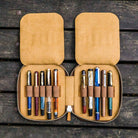 Brown leather pen case with ten pens and zipper on a wooden surface