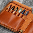 Brown leather zip pen case with writing tools against dark background