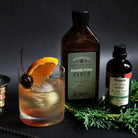 Pine old fashioned cocktail next to pine syrup and pine bitters on dark background