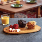 Two muffins on a wooden serving board with fruit and juice, placed on a table.