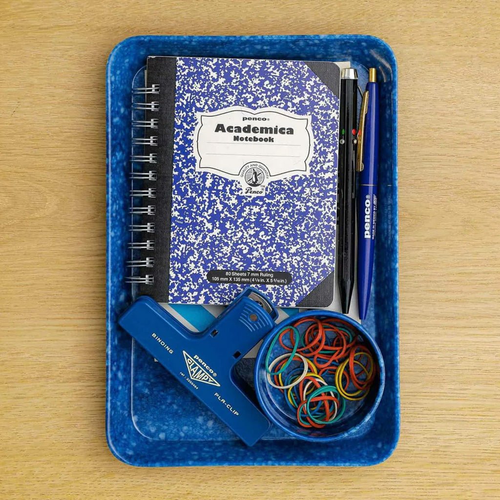 A blue speckled plastic tray holding a blue and white composition notebook, two pens, a blue highlighter, and a small bowl of multicolored rubber bands, arranged on a light wood surface.