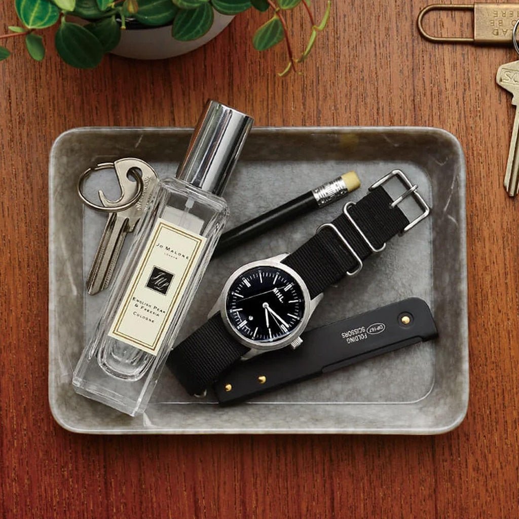 A styled flat-lay on a wooden table showing a clear perfume bottle labeled Jo Malone London, a black watch with a black dial and black fabric strap, a black folding knife, a pencil, and keys arranged in a gray tray.
