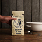 A hand holds a beige paper bag of Marius Fabre Baking Soda on a rustic wooden table, with a white bowl and spoon in the background against a dark wood paneled wall.