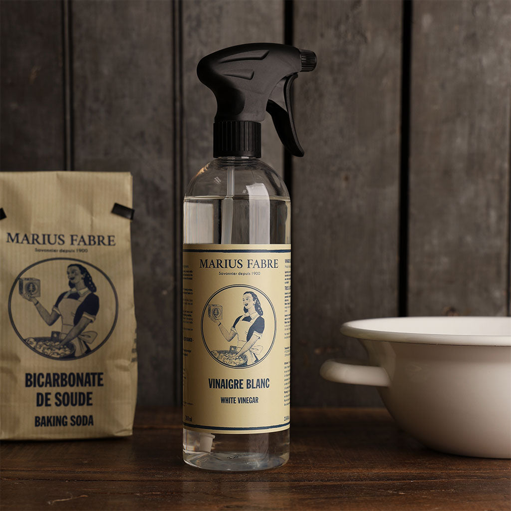 A clear spray bottle of Marius Fabre white vinegar stands on a wooden surface, with a paper bag of Marius Fabre baking soda to the left and a white enamel bowl to the right, all against a dark wood background.