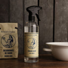 A clear spray bottle of Marius Fabre white vinegar stands on a wooden surface, with a paper bag of Marius Fabre baking soda to the left and a white enamel bowl to the right, all against a dark wood background.
