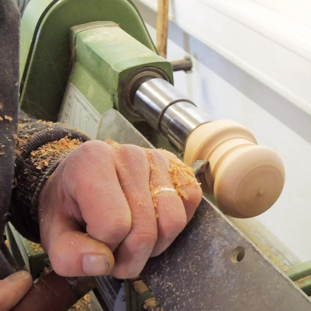 A close-up of a person's hand guiding a woodturning chisel on a wood handle spinning in a lathe machine, with wood shavings flying off.