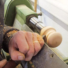 A close-up of a person's hand guiding a woodturning chisel on a wood handle spinning in a lathe machine, with wood shavings flying off.