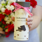 Hand holding tube of deep chocolate caramels with pink ribbon.