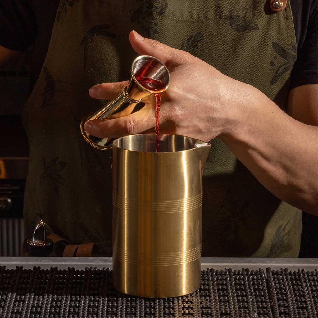 Hand pouring red liquid from cocktail jigger into gold-plated mixtin on bar top