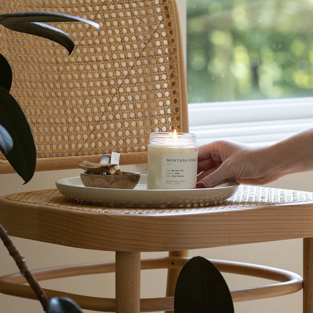 Hand lighting a candle on a small round table with wicker chair and blurred greenery in the background