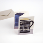 Greeting card with illustrated black-and-cream and blue mugs, displayed upright with a kraft envelope behind.
