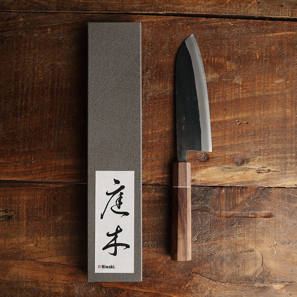A Japanese-style kitchen knife with a dark steel blade and a brown wooden handle, displayed next to its textured gray box.