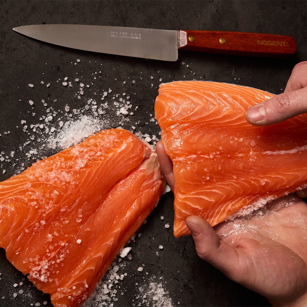 Hands holding salmon and flaky salt next to a wood-handled kitchen knife.