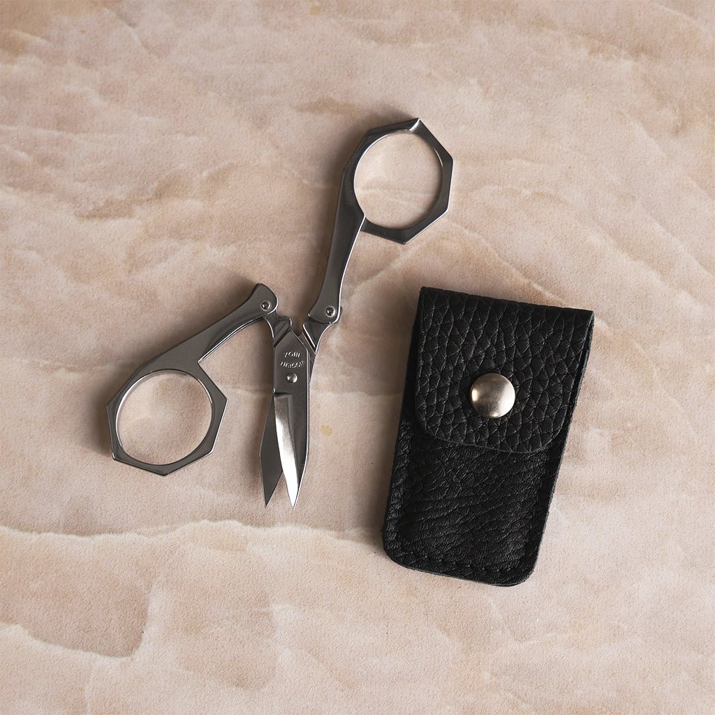 Nogent stainless steel foldable scissors partially open beside a black textured leather case with silver snap closure.
