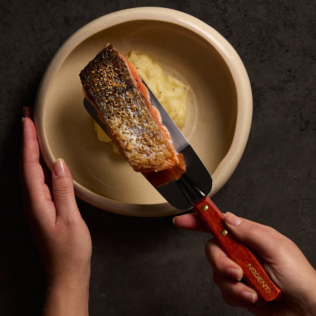Hands holding spatula with piece of salmon above plate of food.