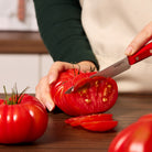 Hand using red-handled tomato knife to slice tomato on wooden surface.