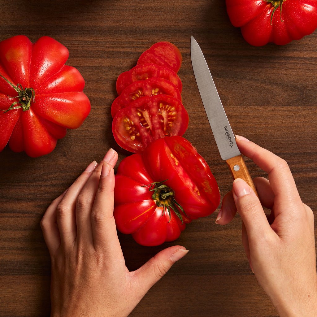 Hand holding knife next to sliced heirloom tomatoes on wooden surface.