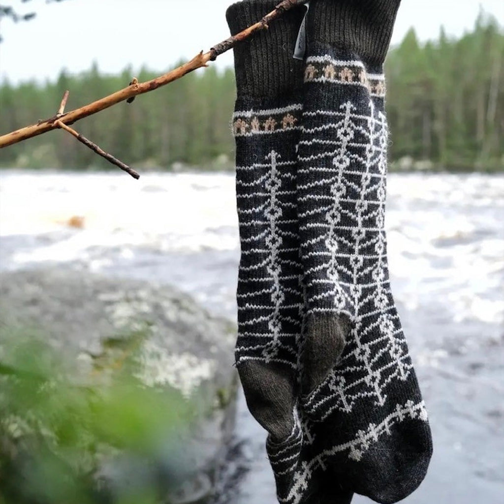 Pair of patterned wool socks hanging on tree branch in front of water setting.