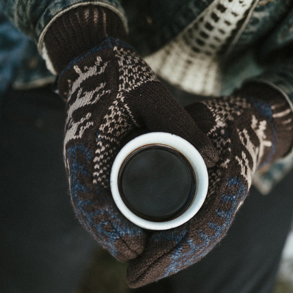 Hands wearing wool mittens and holding mug of coffee.