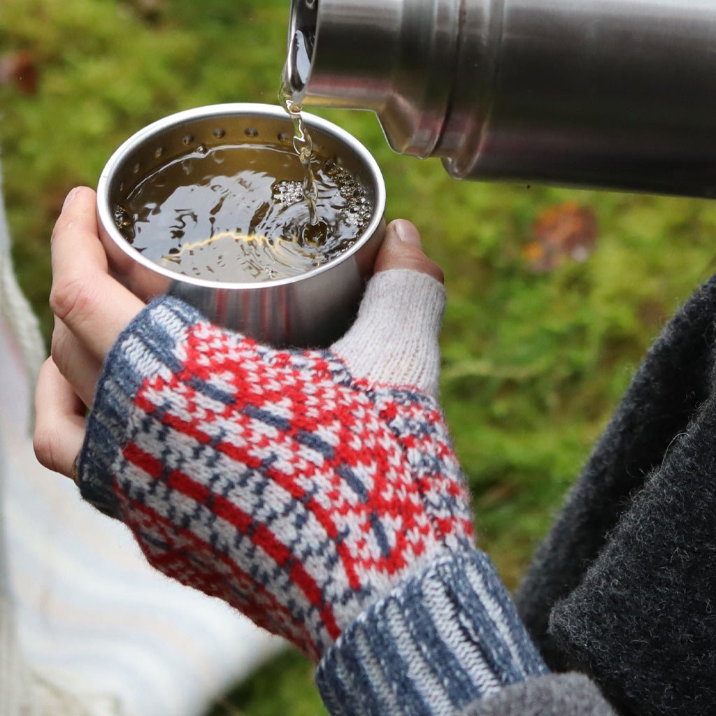 Hands wearing red and blue wool wrist warmers pouring water into metal cup.