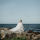 Woman wrapped in patterned white wool blanket in front of water setting.