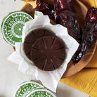 Guajillo chili dark chocolate discs with a brand logo on a wooden board with dried chili peppers.