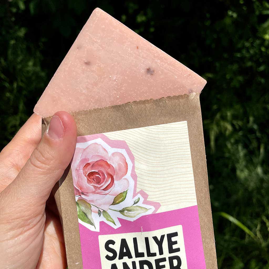 Bar of SallyeAnder organic rose essential soap with a pink rose design on the label, held by a hand against a green leafy background.