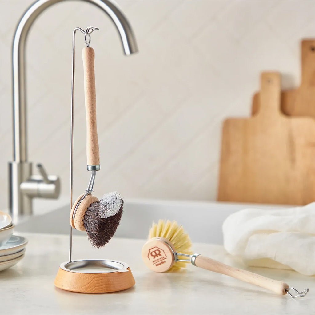A wooden dish brush with dark bristles hanging on hook on kitchen counter next to sink