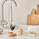A wooden dish brush with dark bristles hanging on hook on kitchen counter next to sink