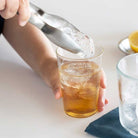 Person adding ice to a glass of iced tea with a lemon wedge on a white surface.