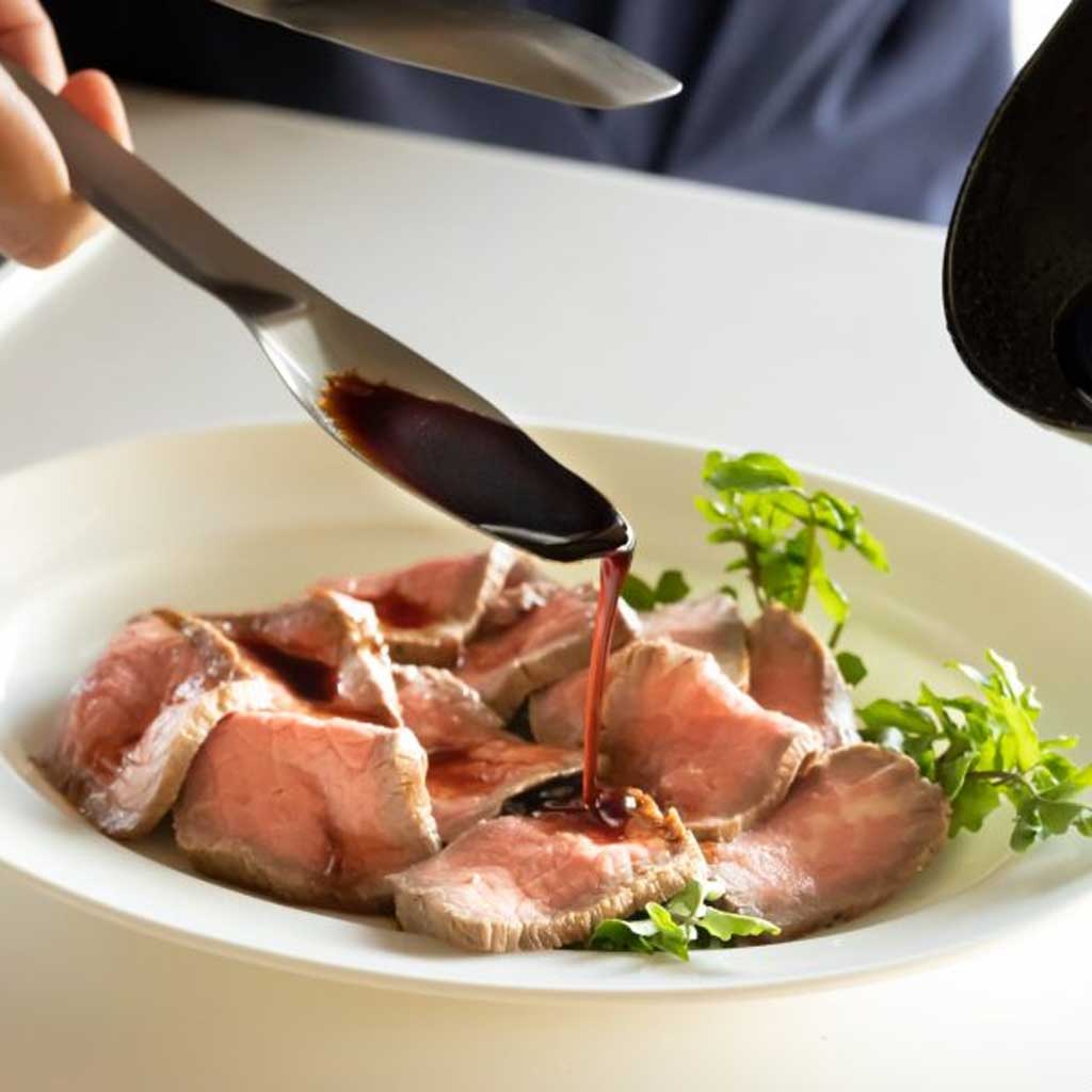 Sliced meat with sauce being poured over it on a white plate.