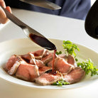 Sliced meat with sauce being poured over it on a white plate.
