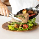 Person using tongs to place a piece of meat onto a salad on a wooden plate.