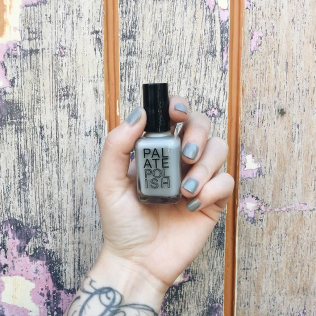 Hand with grey oyster nail polish holding a bottle of 'Palate Polish' nail polish against a textured wall.