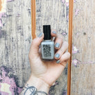 Hand with grey oyster nail polish holding a bottle of 'Palate Polish' nail polish against a textured wall.