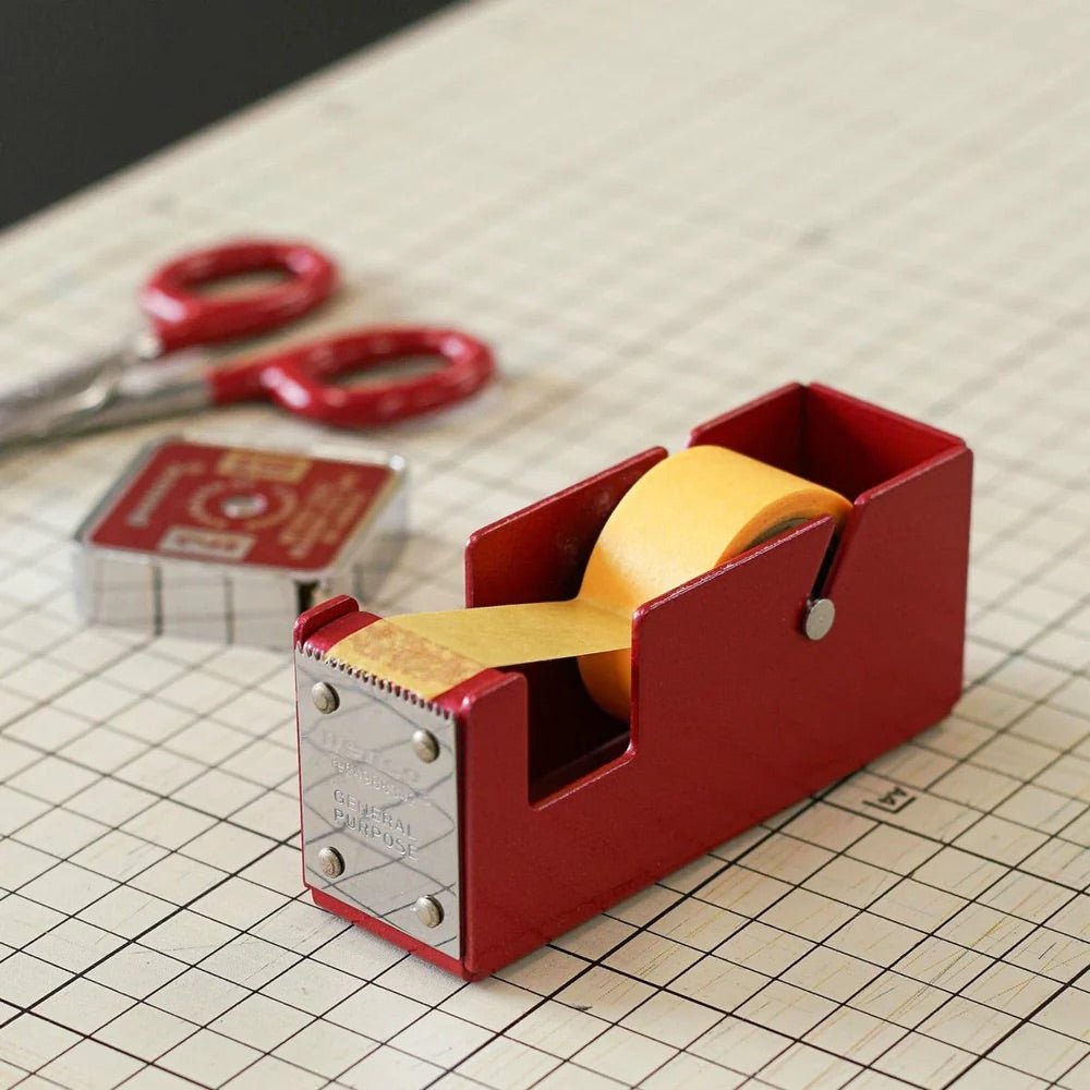 Red tape dispenser with yellow tape on a grid surface with scissors in the background