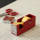 Red tape dispenser with yellow tape on a grid surface with scissors in the background