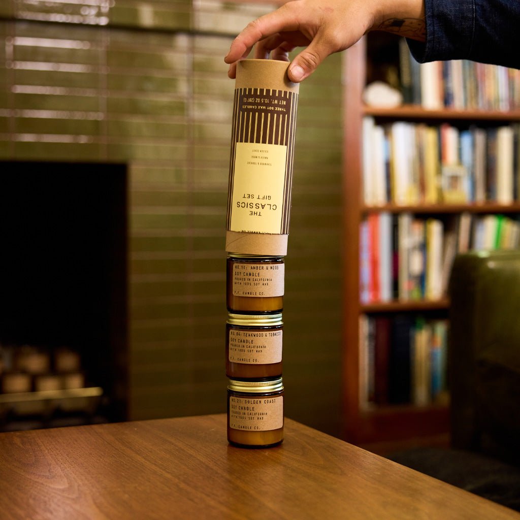 A hand pulling kraft tube with striped label away from stack of three mini candles in amber jars on coffee table in living room