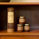 Three P.F. Candle Co. mini candles in amber jars with kraft labels and gold lids next to kraft tube packaging with festive green label on wooden shelf