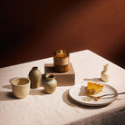 An amber glass jar candles burns on a tablescape set with small vases and a slice of pumpkin pie.